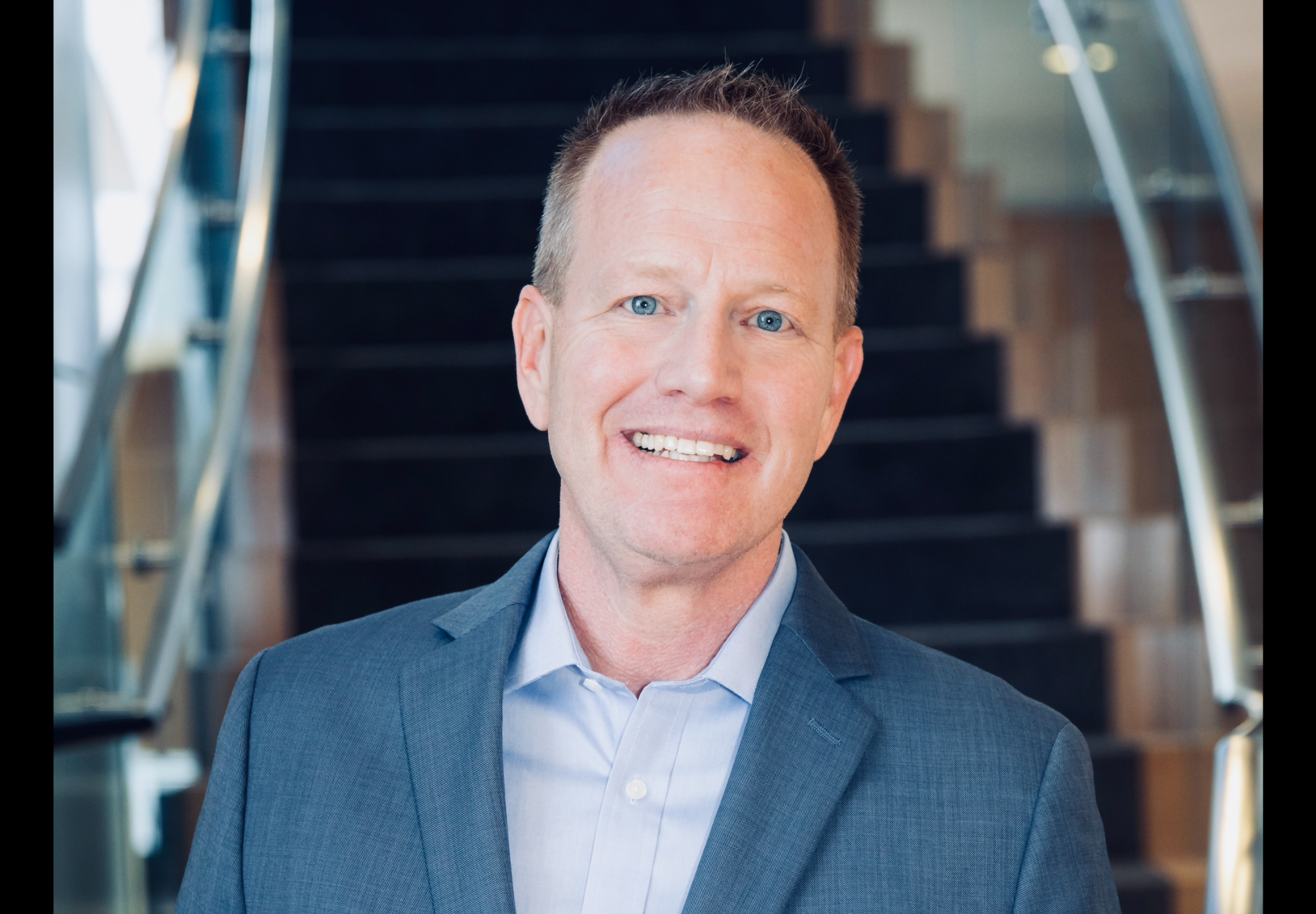 Portrait of Ed Wiley in a blue suit standing in front of a staircase.